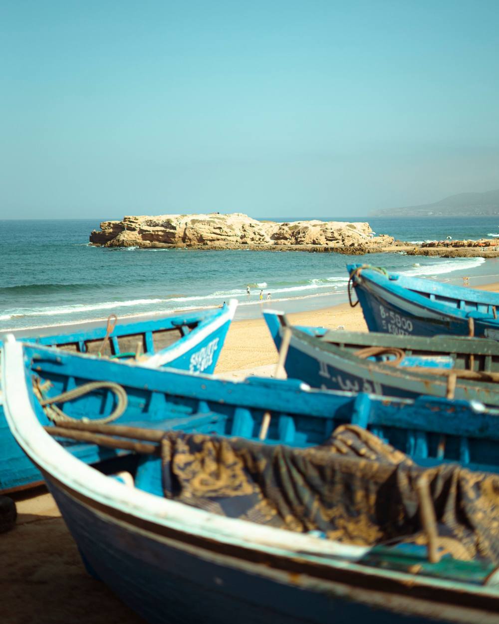 Barques de pêche bleues sur une plage marocaine avec un îlot rocheux au large
